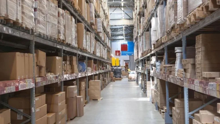 A large warehouse with tall metal racks filled with stacked cardboard boxes and various packaged goods. The warehouse has a high ceiling with industrial lighting, and a wide aisle runs between the storage shelves. In the background, there are additional shelves, some machinery, and a few pallets on the floor.