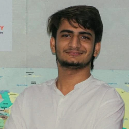 A young man with short brown hair and a trimmed beard, wearing a white shirt, standing in front of a world map with a slight smile.